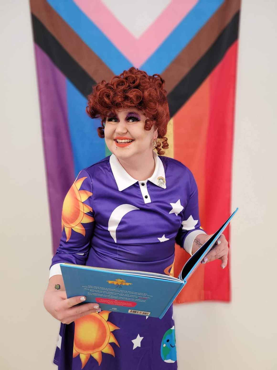 Arya is standing in front of a progress pride flag holding a book and smiling. She is dressed as Ms. Frizzle from The Magic School Bus, with a purple collared dress with the sun, planets and stars on the front.