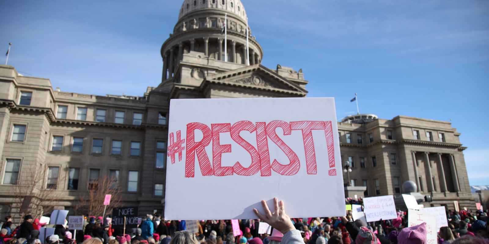 In the center of the image, a person is holding a white sign with "#Resists" in red letters there is a crowd of people in front  of the Idaho state capitol building in the background