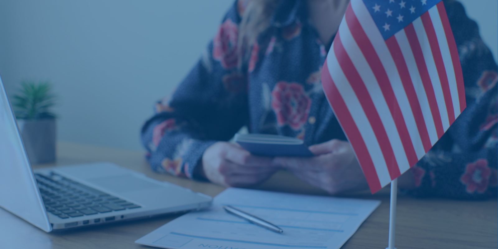 Picture of woman sitting at a table with a passport in her hands, face out of frame. There is a laptop, paper and pen, and small American flag on the table in front of her.