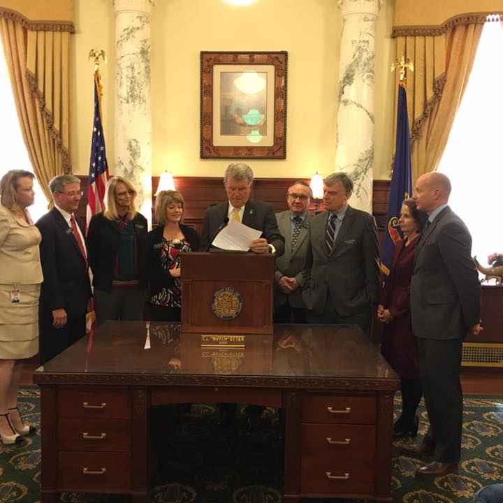 Gov. Otter standing at a podium signing a bill into law in the Governor's office with people standing around him
