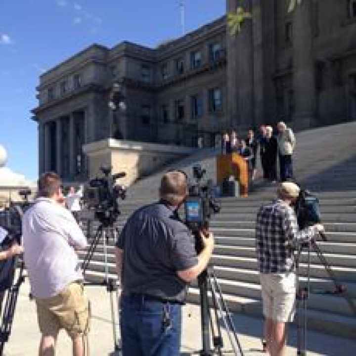 Press conference in front of the Idaho Statehouse. cameras in the fore ground, people beyond a podium in the background