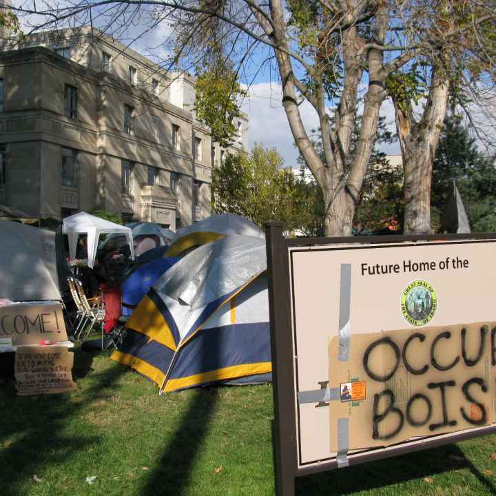 a sign with a cardboard sign taped on top with spray painted "occupy boise" on it with tents in the background