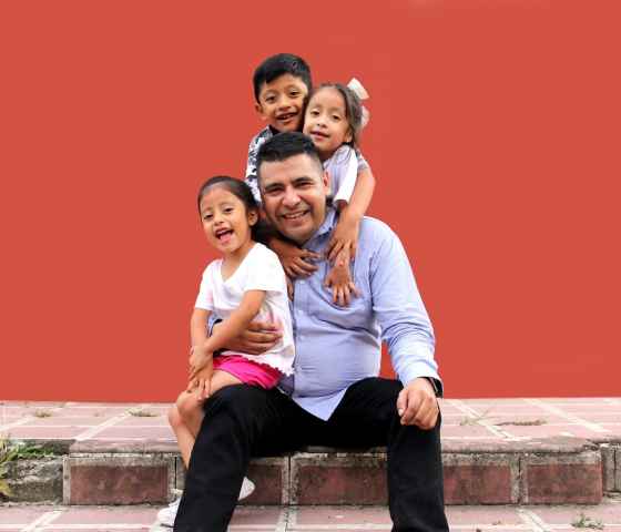 A Mexican father sits on a stoop smiling as a 4-year-old daughter sits on his right lap, her twin sister hugs the father from behind, and an 8-year-old boy embraces them all from behind. All family members are smiling in front of a terra cotta background.